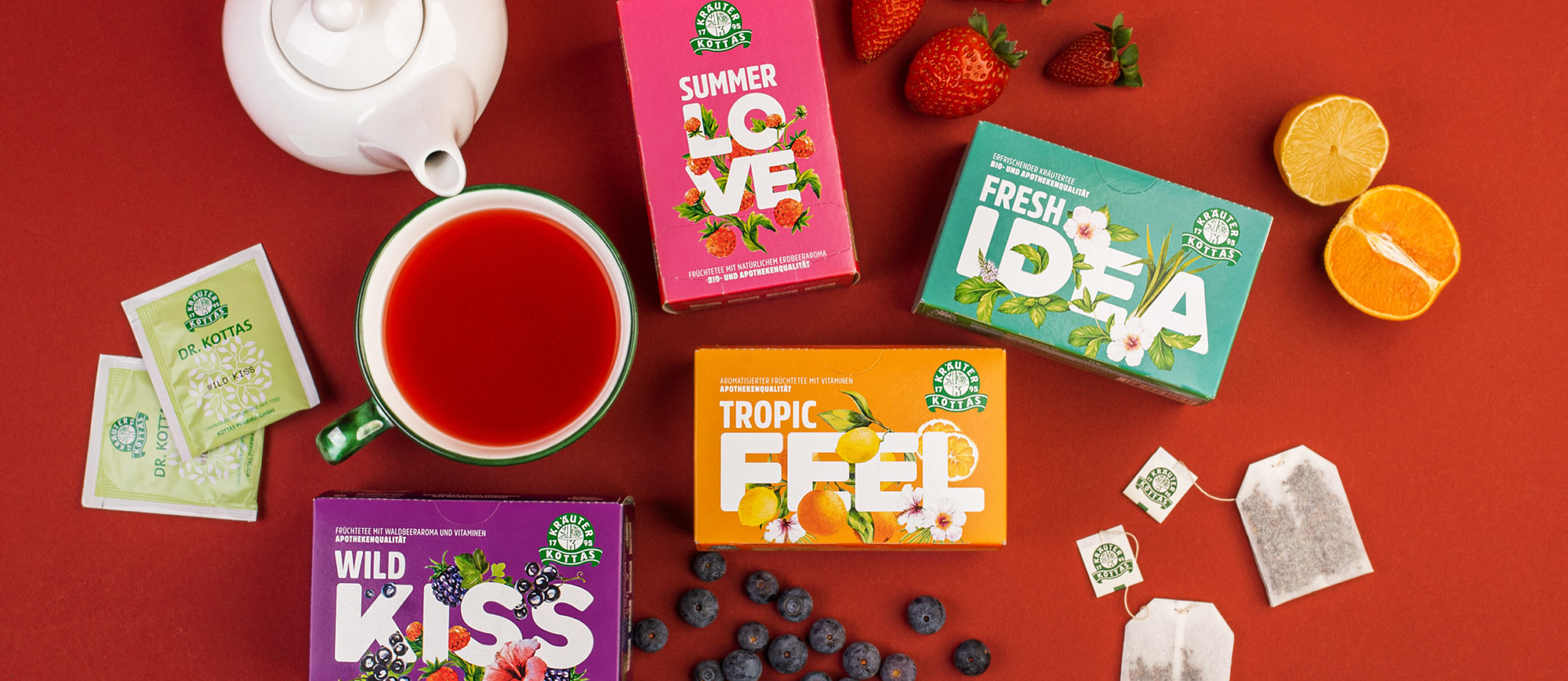 Teekanne, Tasse mit Tee, fünf Teepackungen, Erdbeeren, Blaubeeren und eine halbierte Orange auf einem Tisch.