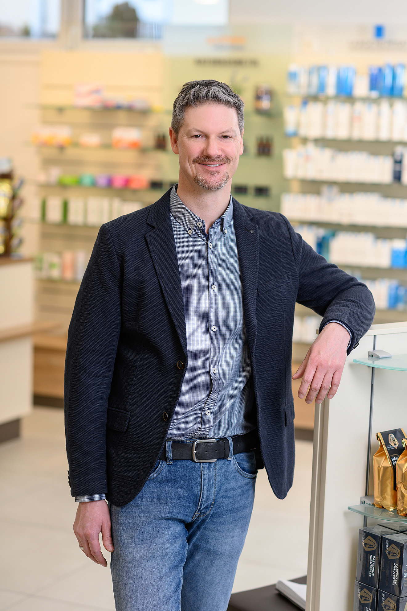 Apotheken Inhaber Stefan Gritsch Stefan Gritsch - Inhaber und Apotheker in Sakko und Jeans steht entspannt in seiner hellen Apotheke und lächelt freundlich in die Kamera.