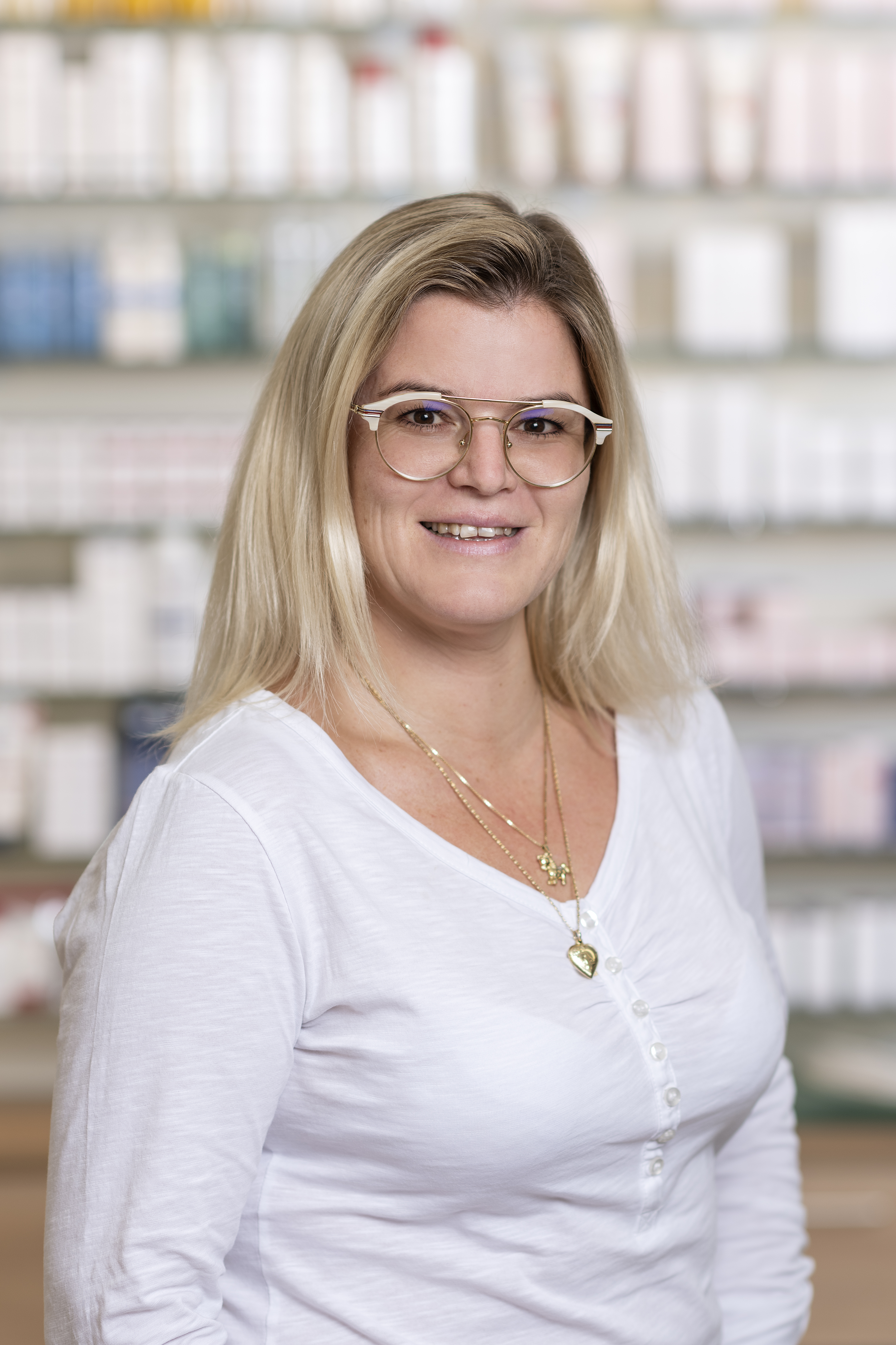 Frau mit mittellangem Haar und Brille lächelt freundlich. Sie trägt ein Oberteil mit Knopfleiste sowie mehrere Ketten. Im Hintergrund sind unscharfe Apothekenregale zu sehen.