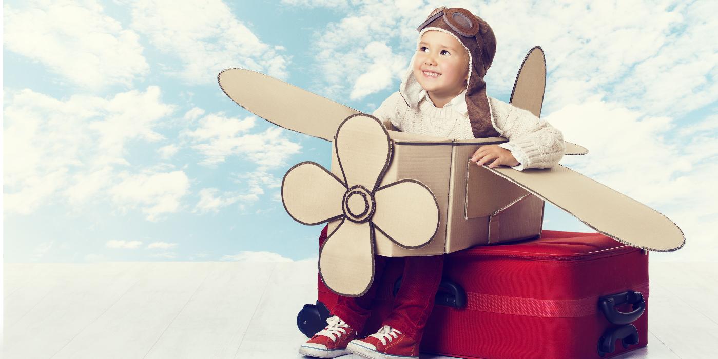 Urlaubsstimmung und Fantasie Kind im Pappflugzeug-Kostüm auf rotem Koffer vor Himmelshintergrund – Urlaubsstimmung und Fantasie.