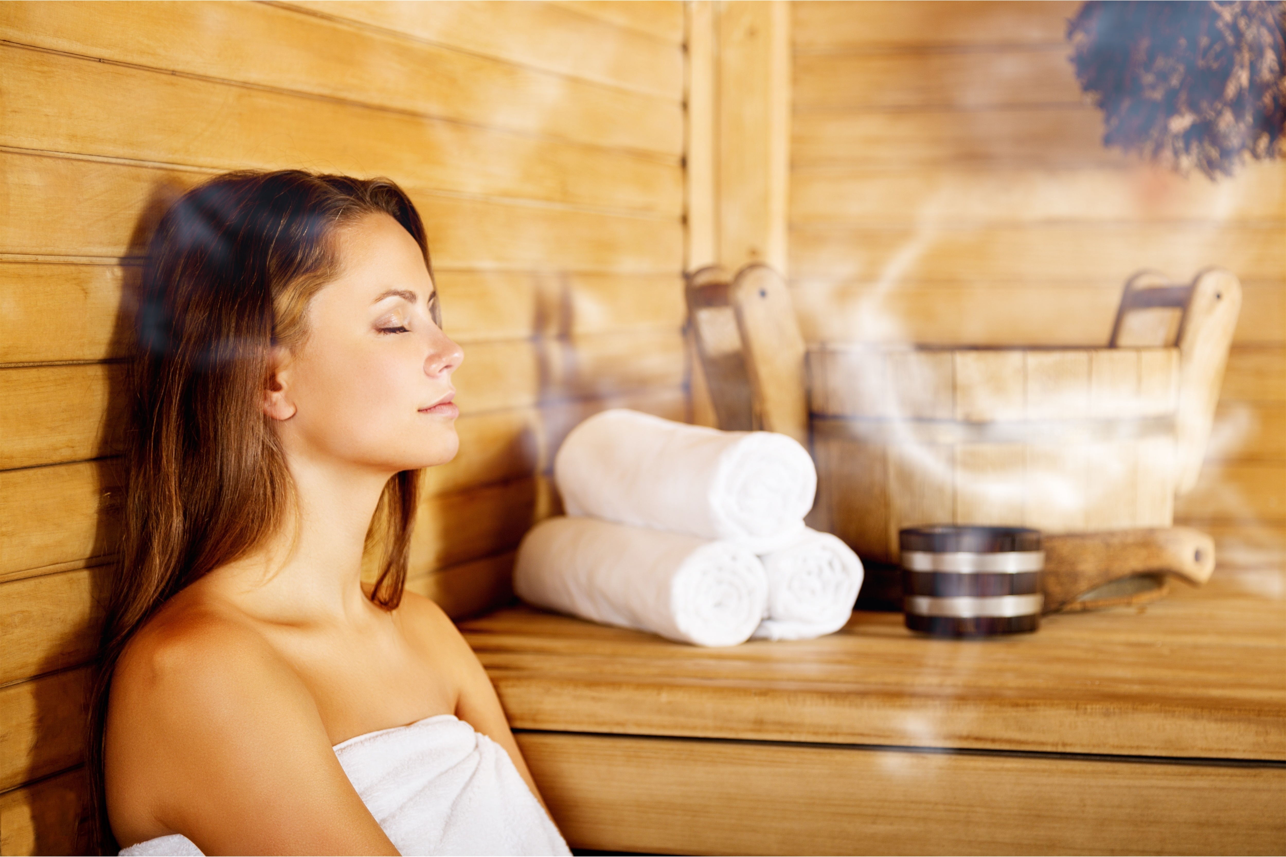 In der Sauna Frau entspannt in der Sauna mit geschlossenen Augen, umgeben von Dampf, Holzinterieur und gerollten Handtüchern.