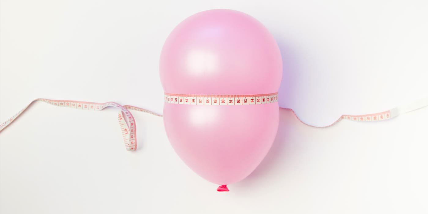 Blähbauch loswerden Ein aufgeblasener Ballon wird von einem Maßband umschlossen – symbolisiert einen aufgeblähten Bauch oder Körperumfang.