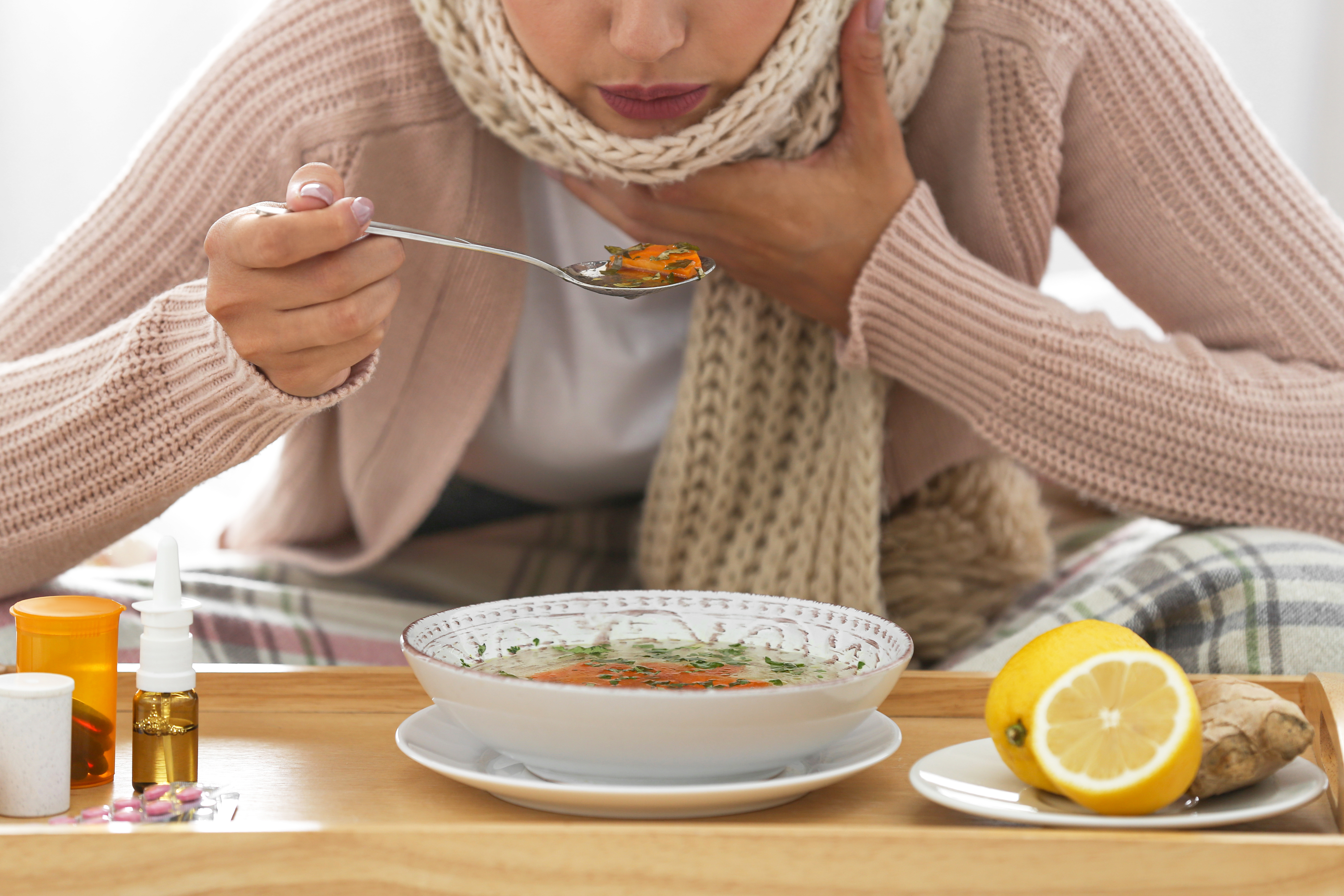 Erkältung versorgen Erkältete Person isst Suppe, hält sich den Hals – daneben Hausmittel und Medikamente auf einem Tablett.