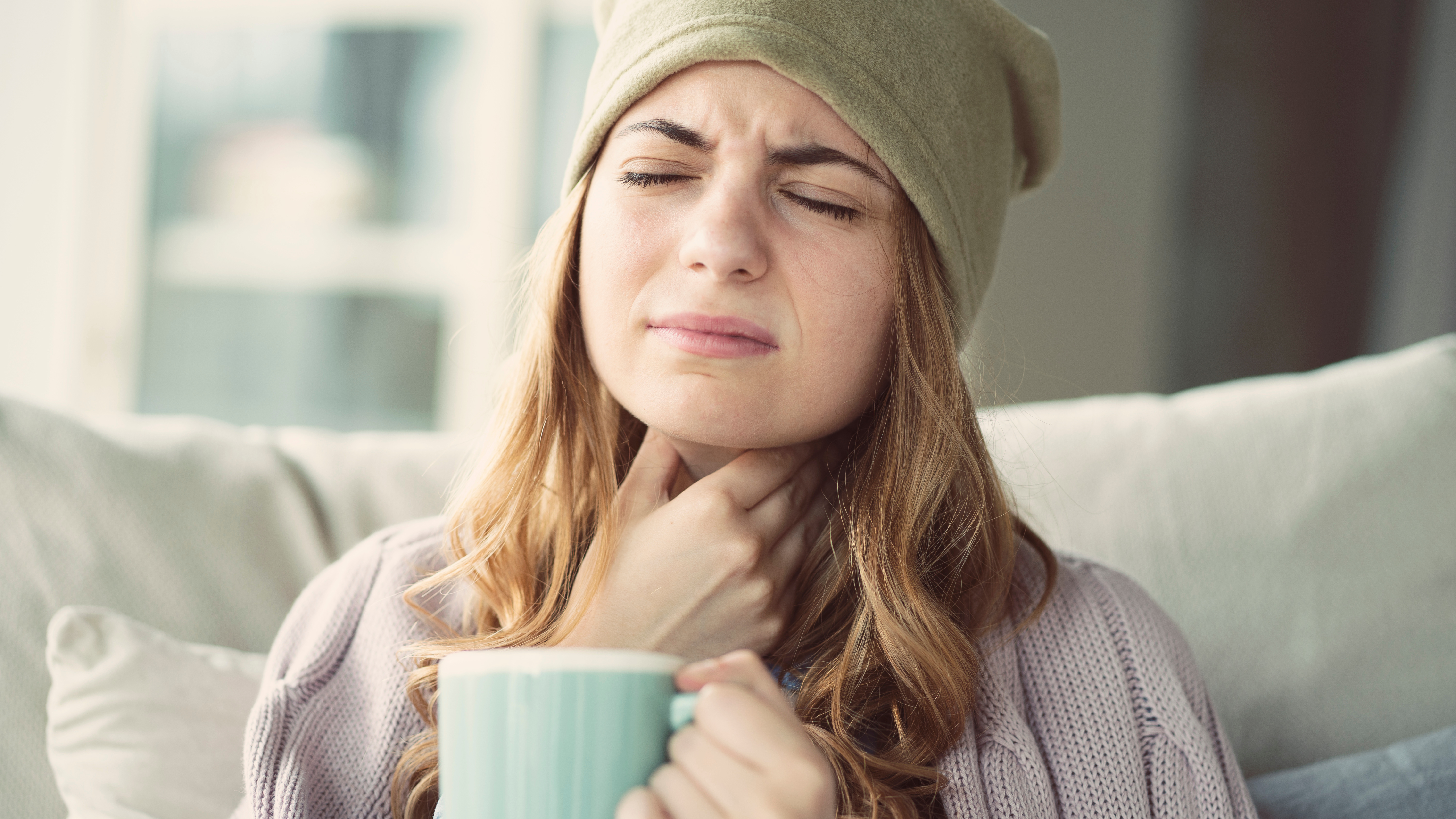 Halsscherzen lindern Junge Frau mit Halsschmerzen hält sich den Hals und trinkt aus einer Tasse, Blick schmerzverzerrt, trägt Mütze und Strickpullover.