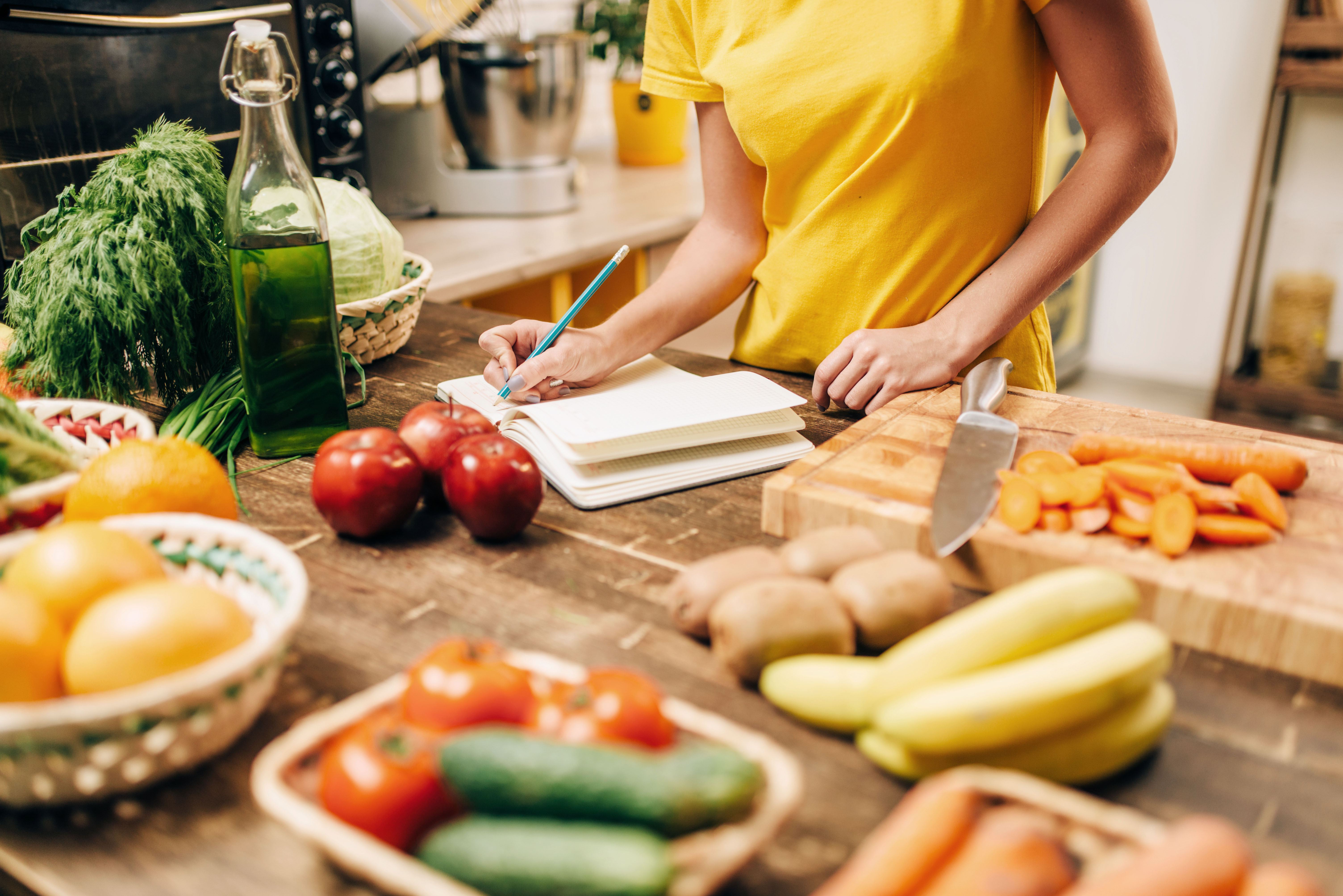 Gesunde Ernährung Person schreibt Rezept auf, umgeben von frischem Gemüse und Obst in einer Küche – gesunde Ernährung planen.